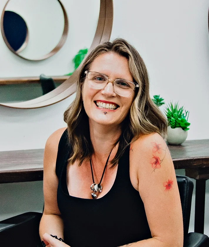 Nadine, Stylist at Hair Management Co in Gymea, smiling in the salon with wavy highlighted hair and glasses